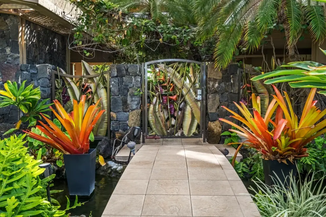 A tiled garden path leads to a gate framed by tropical plants and dark stone walls.