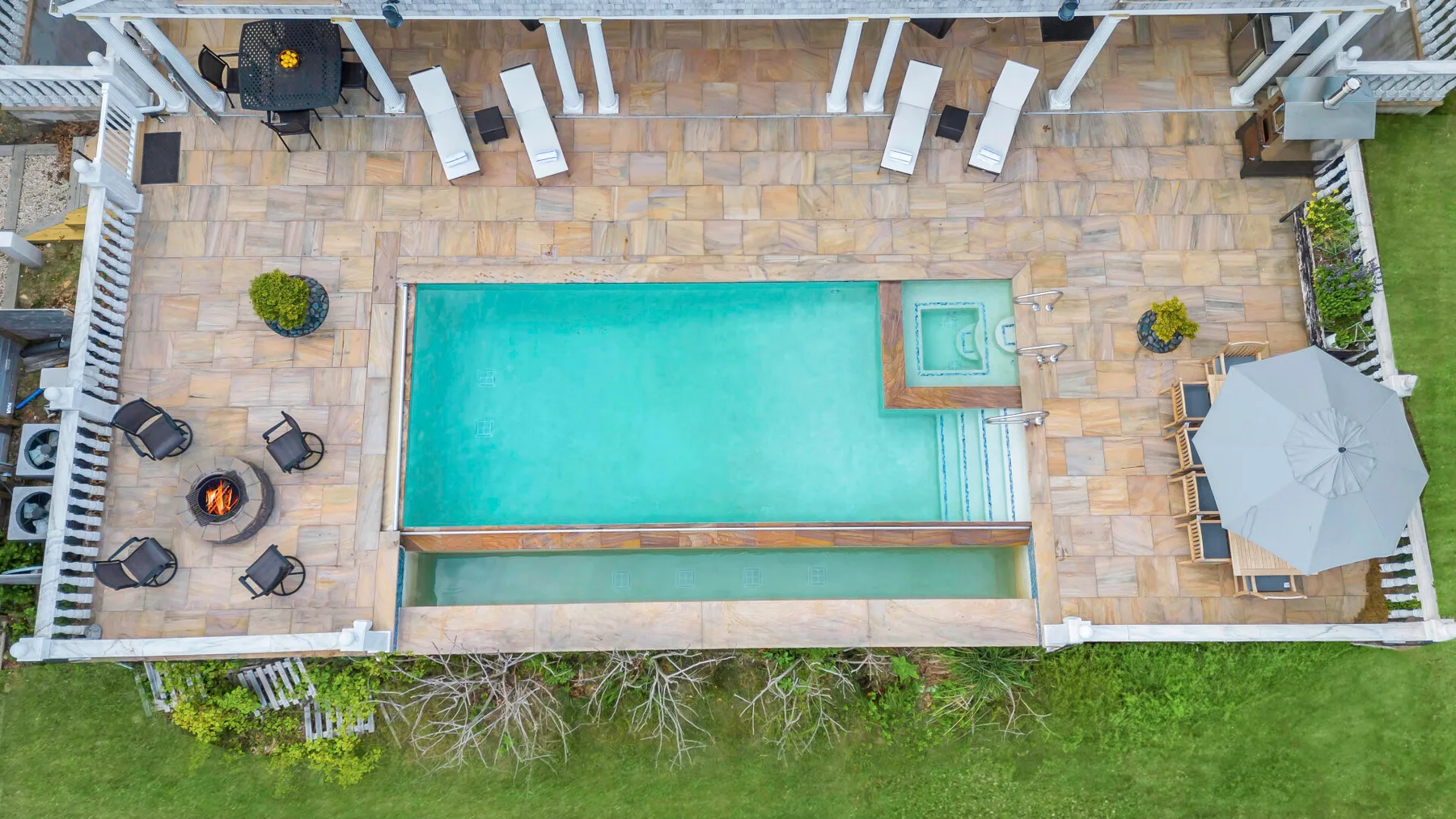 A swimming pool sits on a paved patio with outdoor seating and an umbrella.