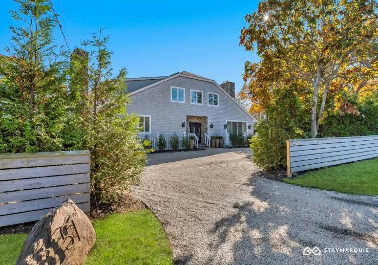A gray house sits beyond a gravel driveway, framed by trees and white fencing.