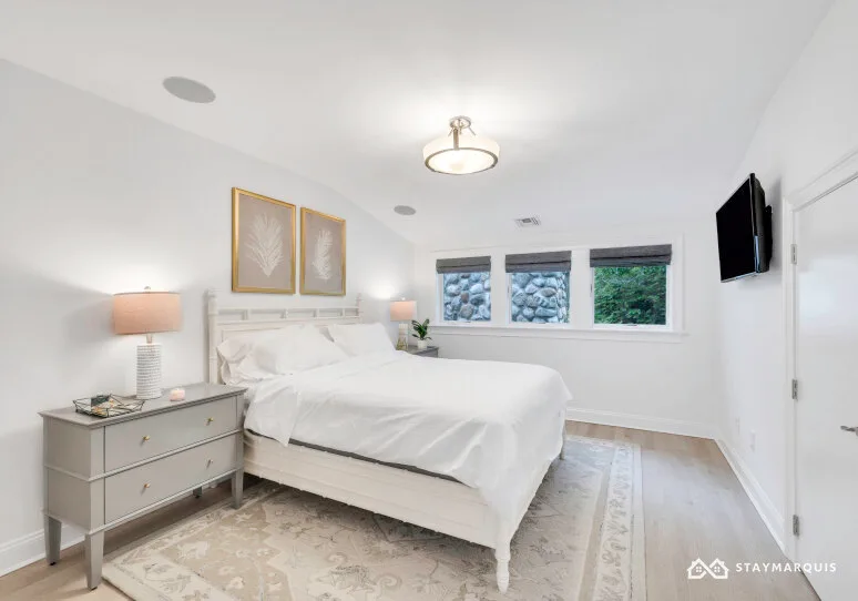 A bright bedroom with a neatly made bed and windows along the far wall.