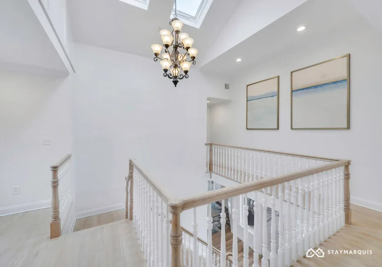 A bright upstairs landing has white railings, framed artwork, and a chandelier.