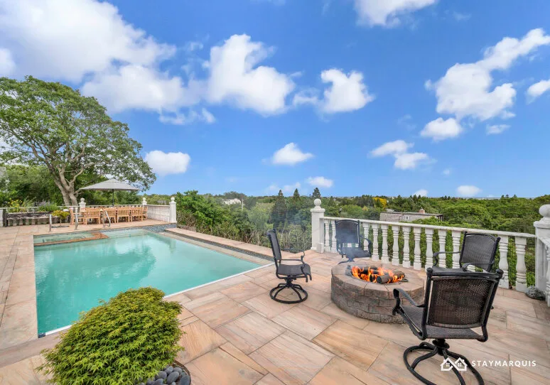 A sunny outdoor pool terrace features a seating area and a round fire pit.