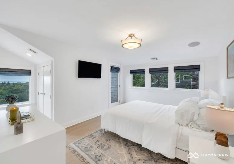 A bright bedroom with a white bed, several windows, and a television.