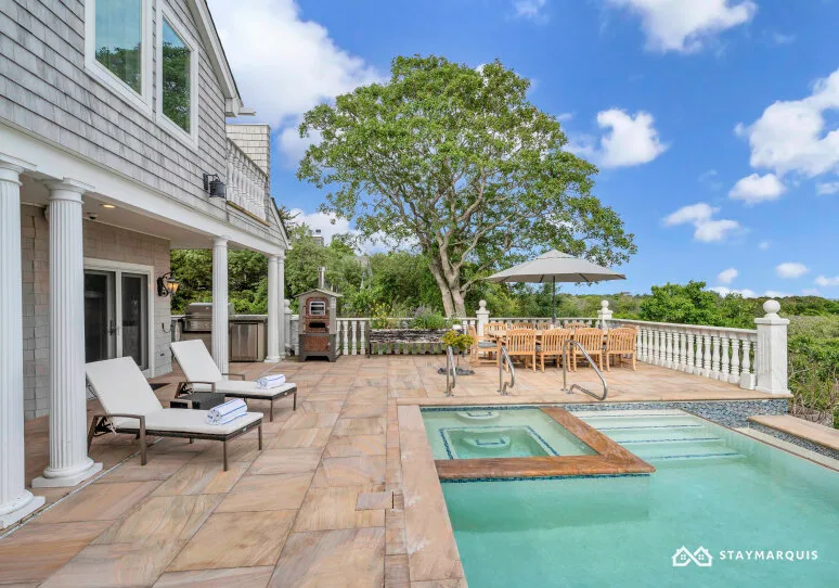 A paved outdoor pool area with lounge seating, a spa, and a shaded dining table.