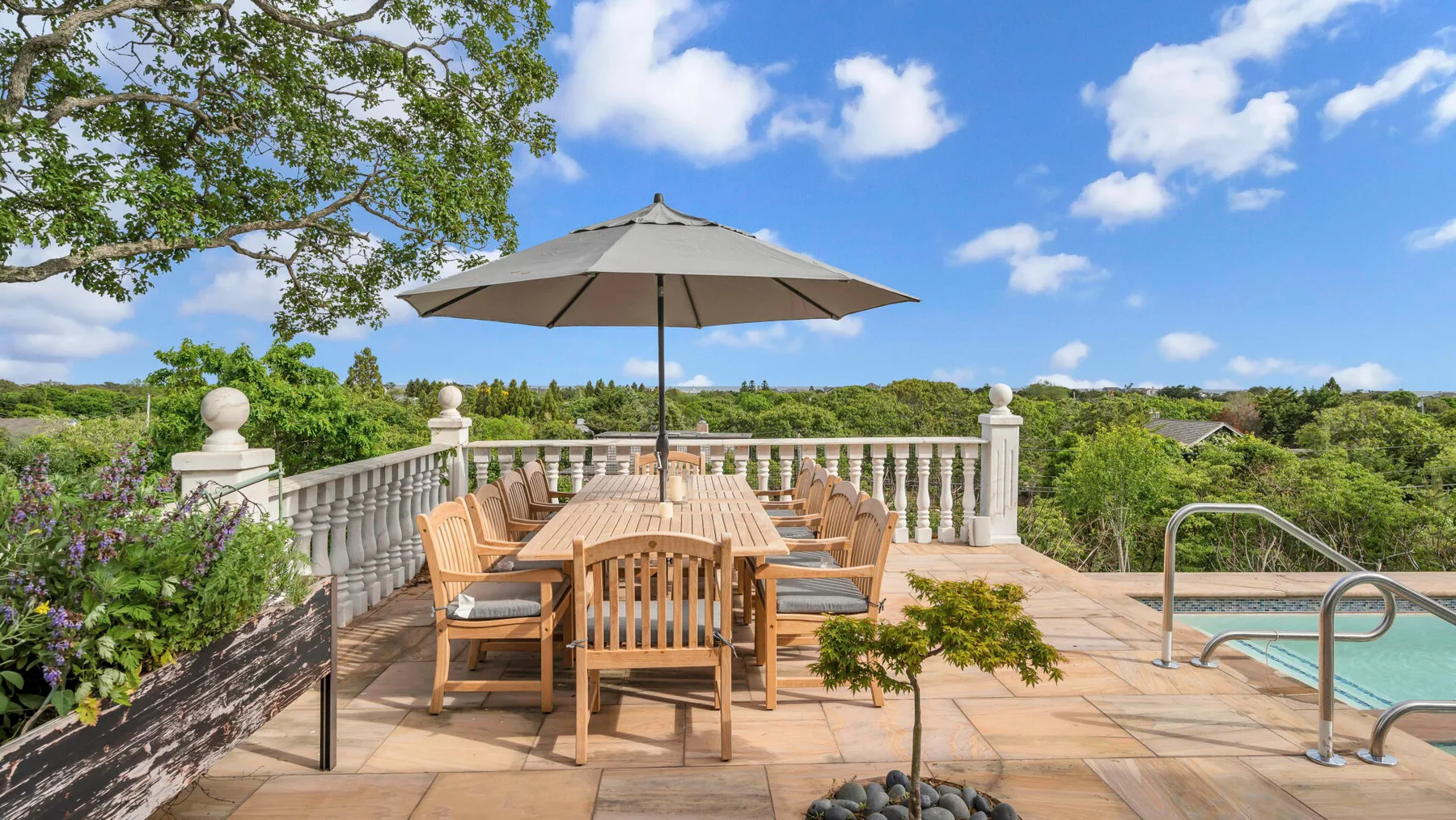 A terrace dining area with wooden seating and a large umbrella overlooks a swimming pool.