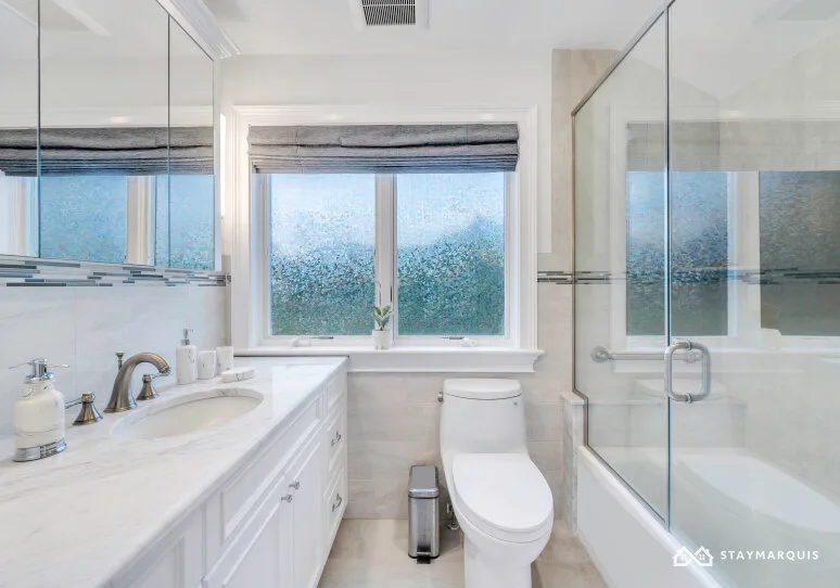 A bright bathroom with a vanity, toilet, and glass shower beside a window.