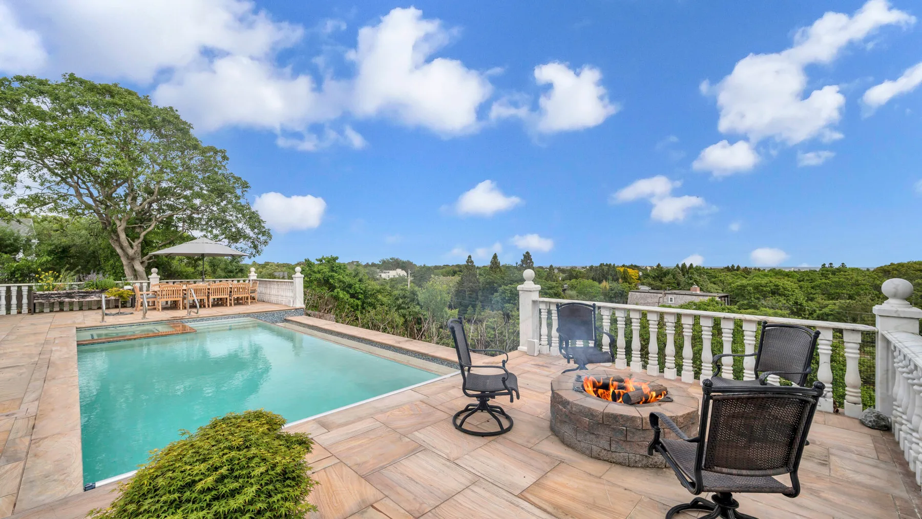 A stone patio with a swimming pool, seating, and a fire pit overlooks green countryside.