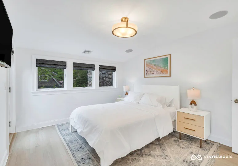 A bright bedroom with a white bed, windows, and a bedside table.