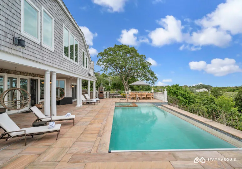 An outdoor swimming pool beside a house, with a paved terrace and lounge seating.