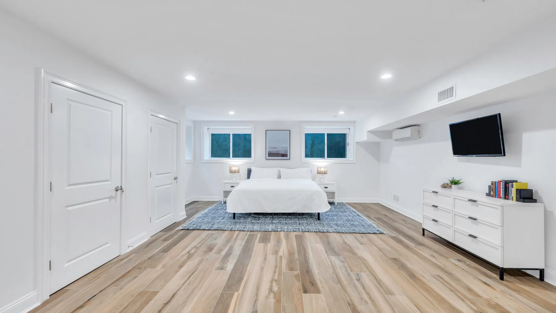 A bright bedroom with a bed, wood flooring, and white built-in storage.