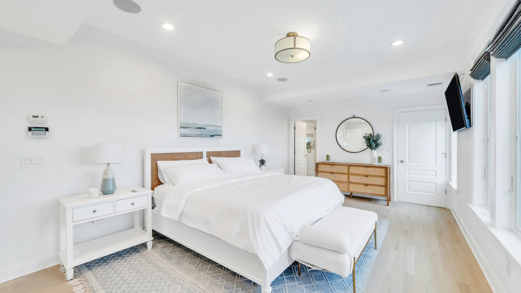 A bright bedroom with a neatly made bed and a dresser beneath a round mirror.
