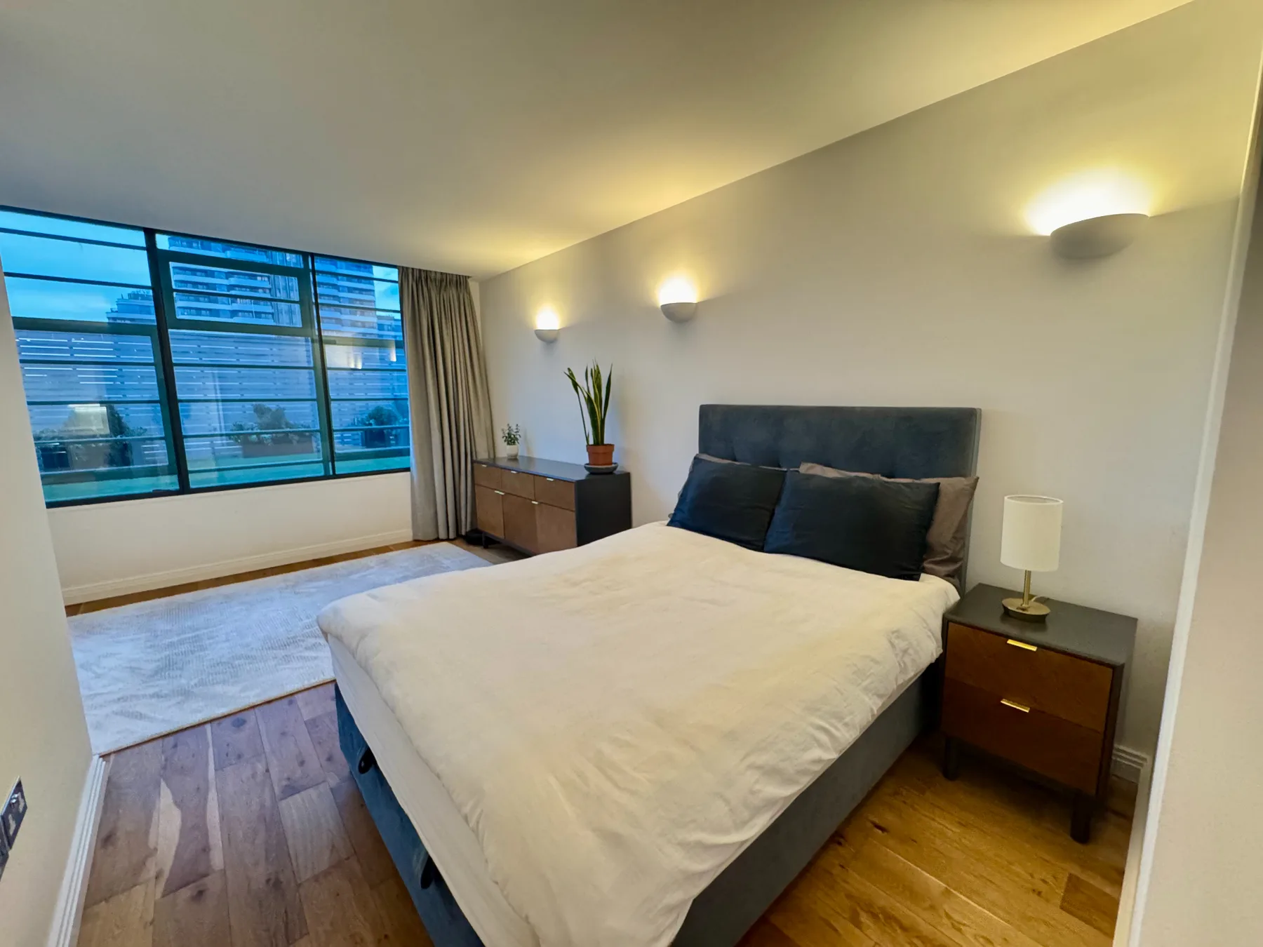 Un dormitorio con una cama junto a una ventana amplia y suelo de madera oscura.