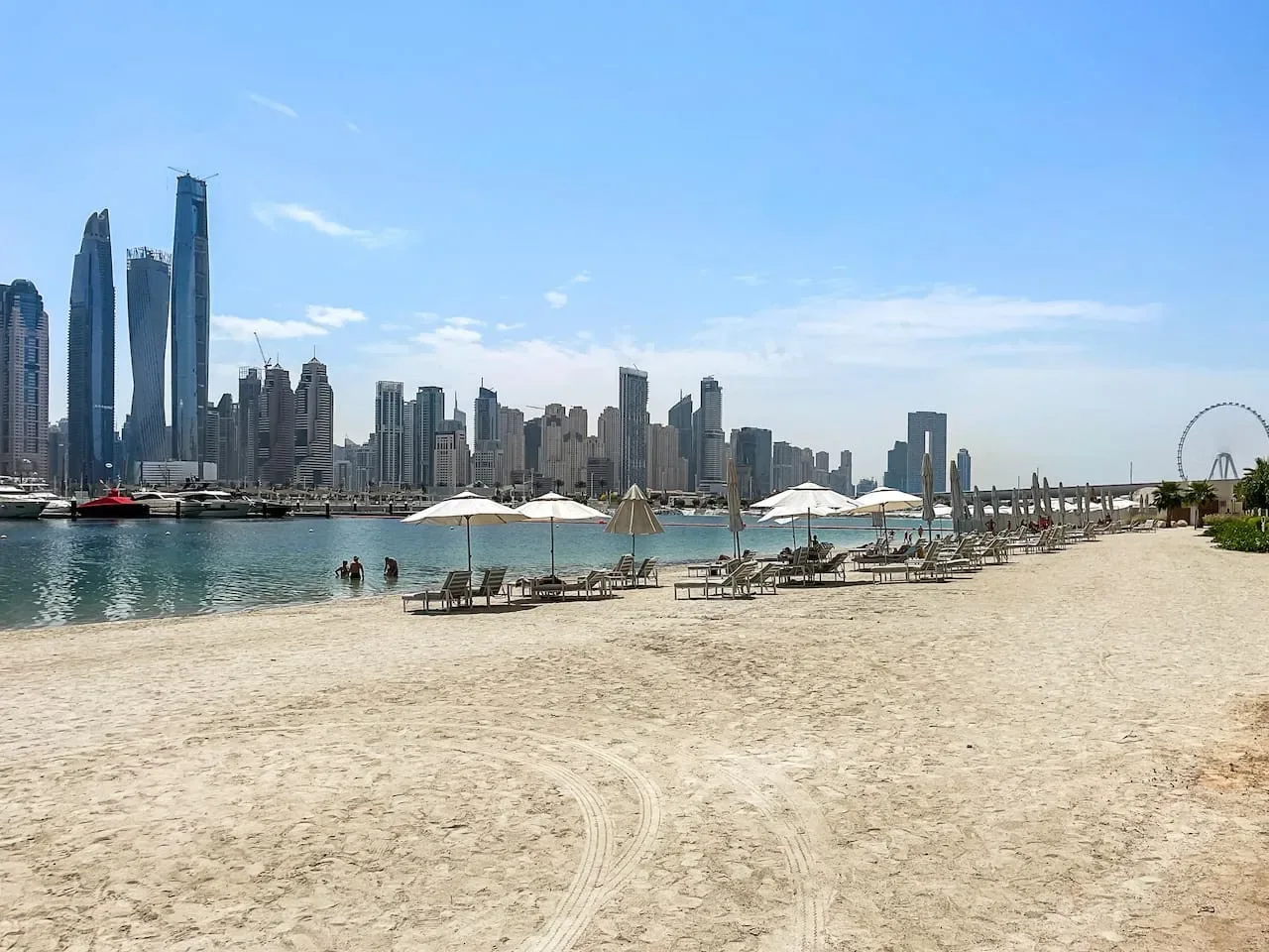 Una playa de arena junto al agua, con asientos y un horizonte urbano al otro lado.