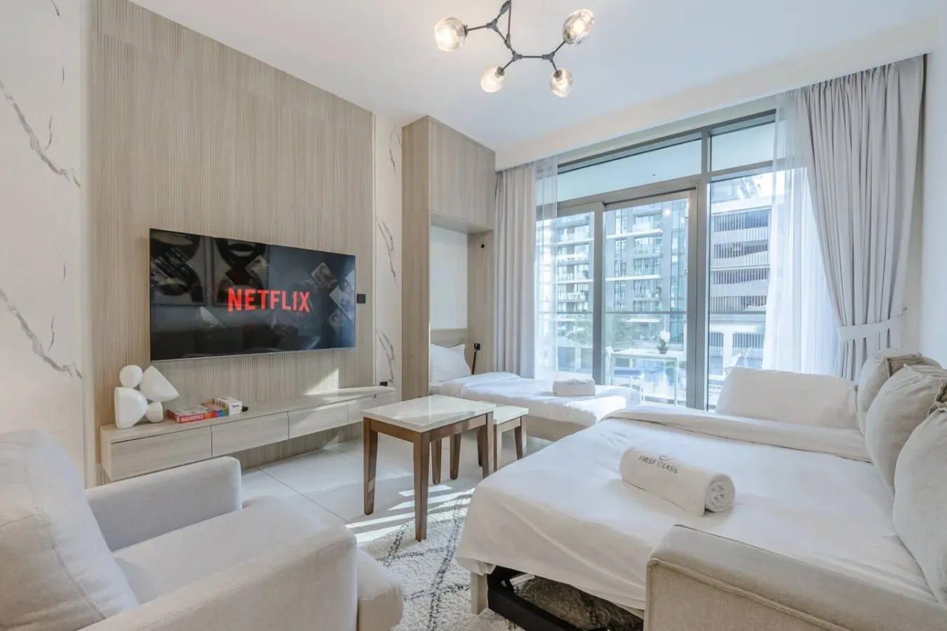 Una luminosa sala de estar tiene asientos blancos, un televisor y grandes ventanales.