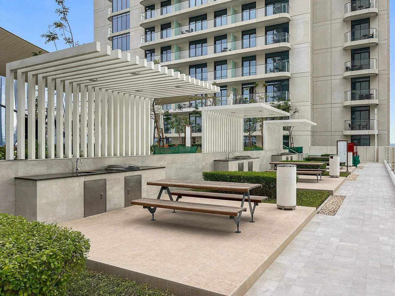 Un patio exterior tiene mesas de picnic bajo pérgolas blancas, con un edificio de apartamentos al fondo.