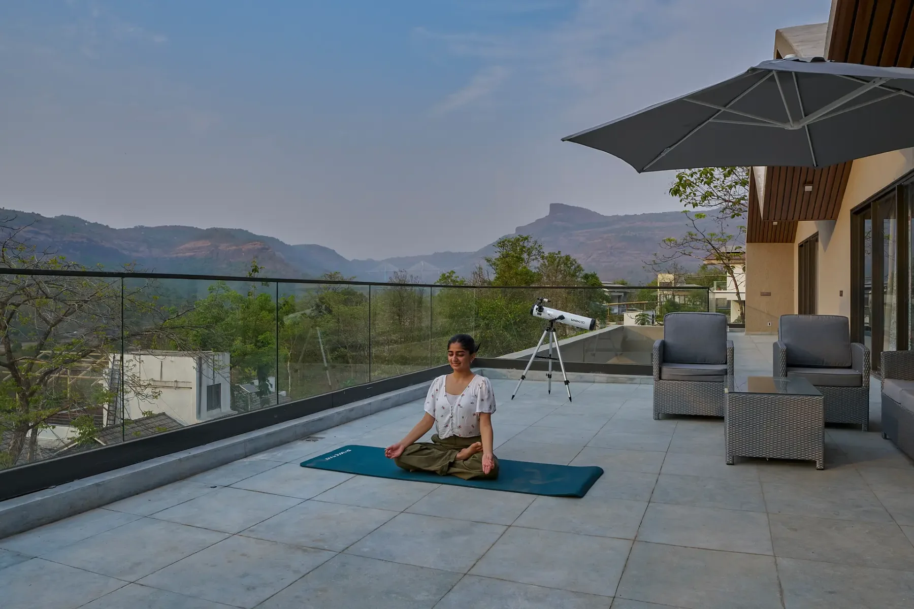 Una persona está sentada en una esterilla de yoga en una terraza con vistas a las montañas.