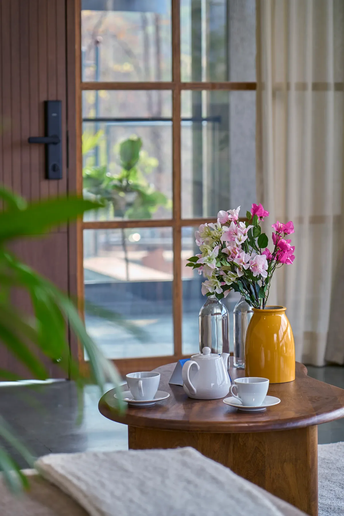 Una mesa de té con tazas y flores está junto a una ventana grande.