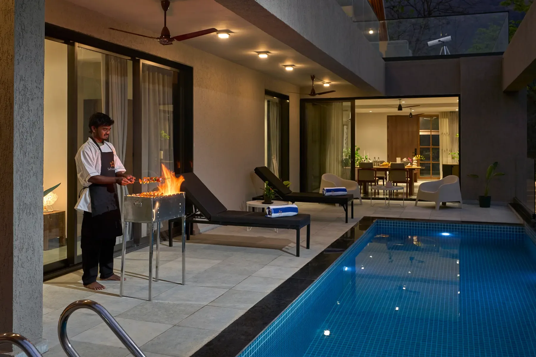 Un patio cubierto junto a una piscina, con una persona cocinando en una parrilla exterior.
