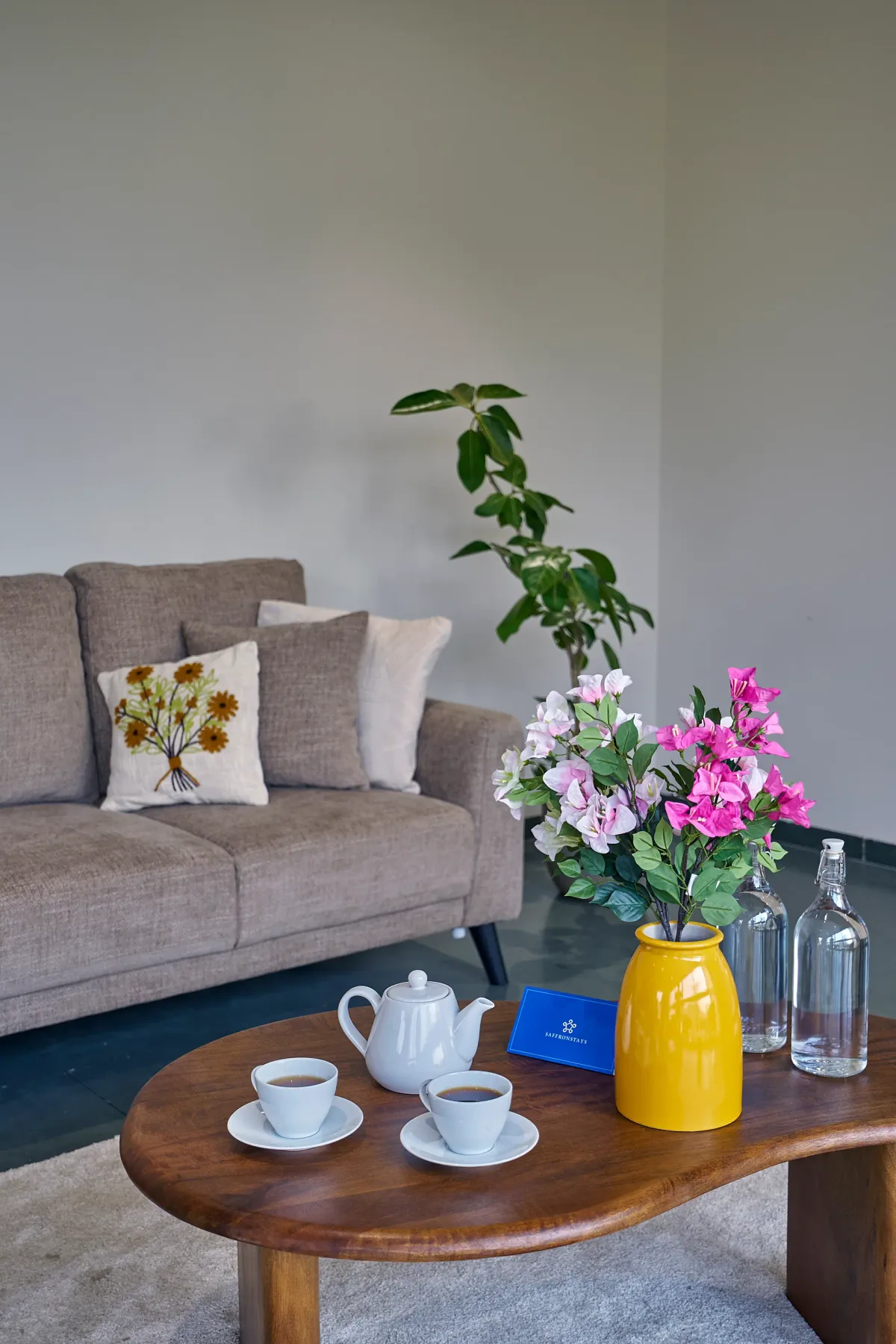 Una sala de estar con un sofá gris, plantas con flores y una mesa de centro de madera.