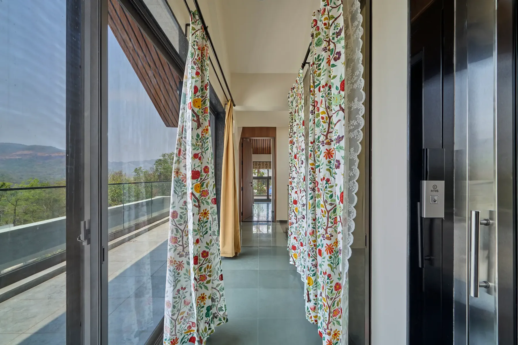 Un pasillo luminoso con cortinas florales y puertas de cristal hacia un balcón.