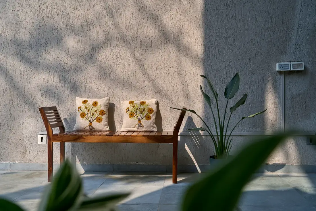 Un banco de madera al aire libre con cojines florales está junto a plantas frondosas.