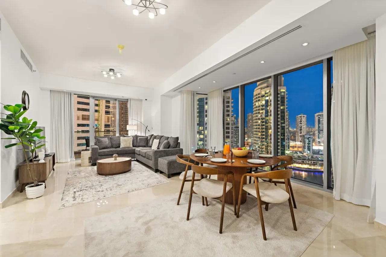 Una luminosa sala de estar y comedor con asientos, una mesa y amplias ventanas con vistas a edificios de la ciudad.