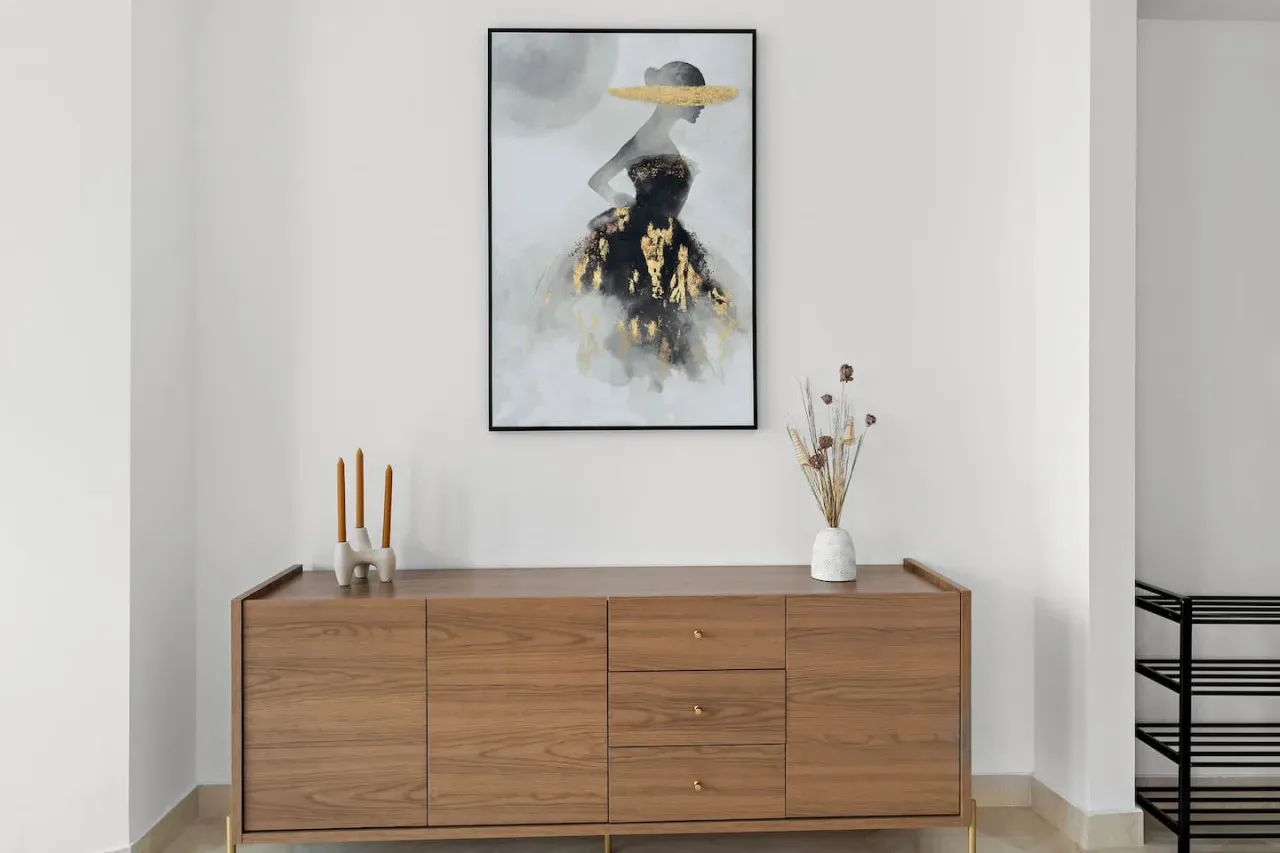 Un aparador de madera está bajo una obra enmarcada en una habitación luminosa y sencilla.