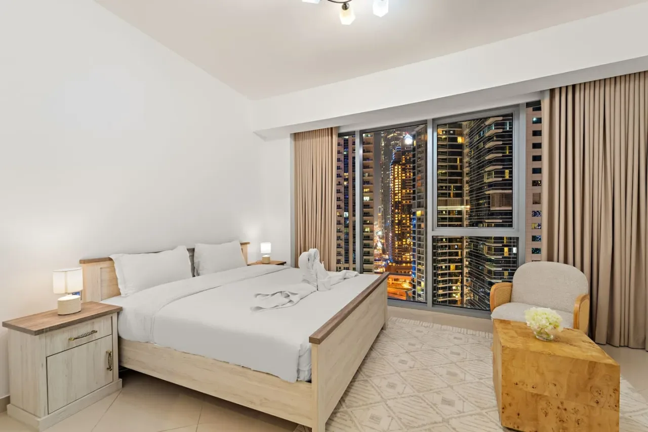 Un dormitorio con una cama bien tendida junto a grandes ventanales con vistas a edificios de la ciudad.