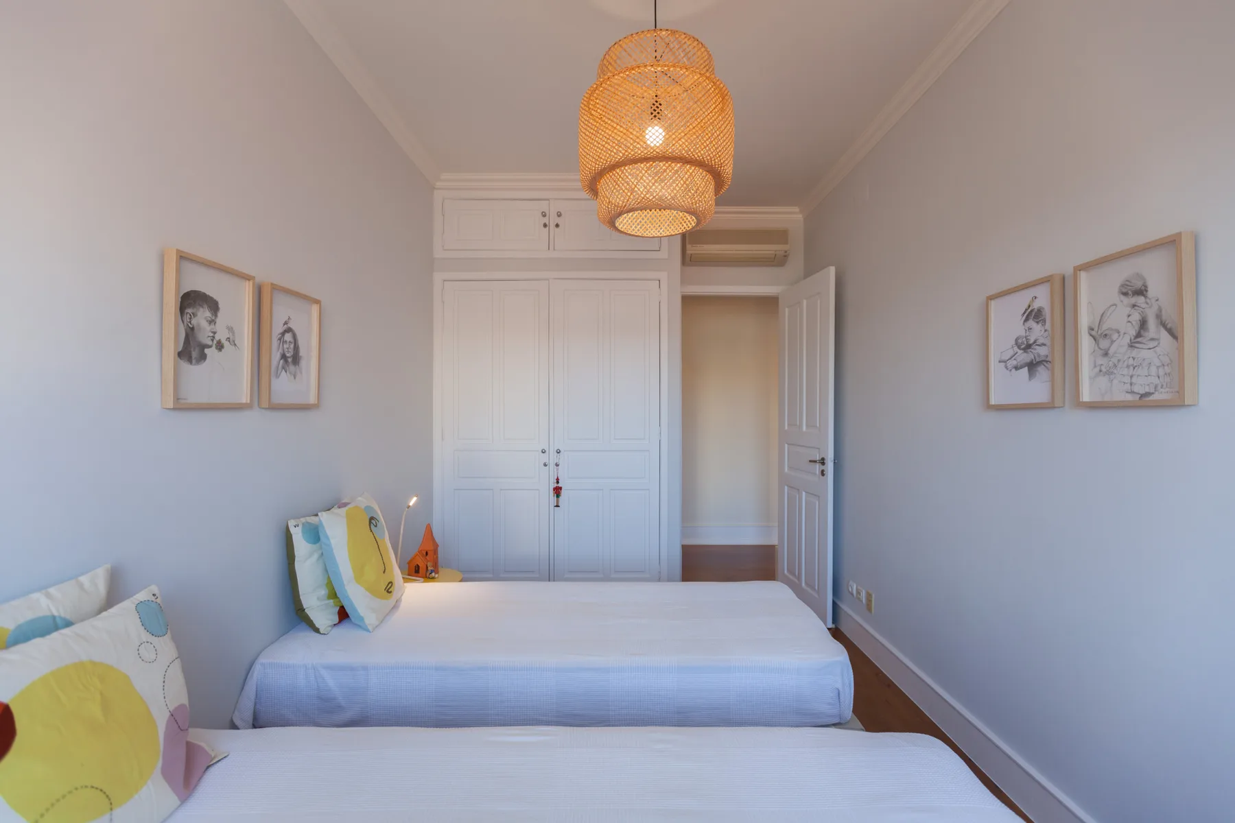 A bright bedroom with two beds, framed artwork, and a woven pendant light.