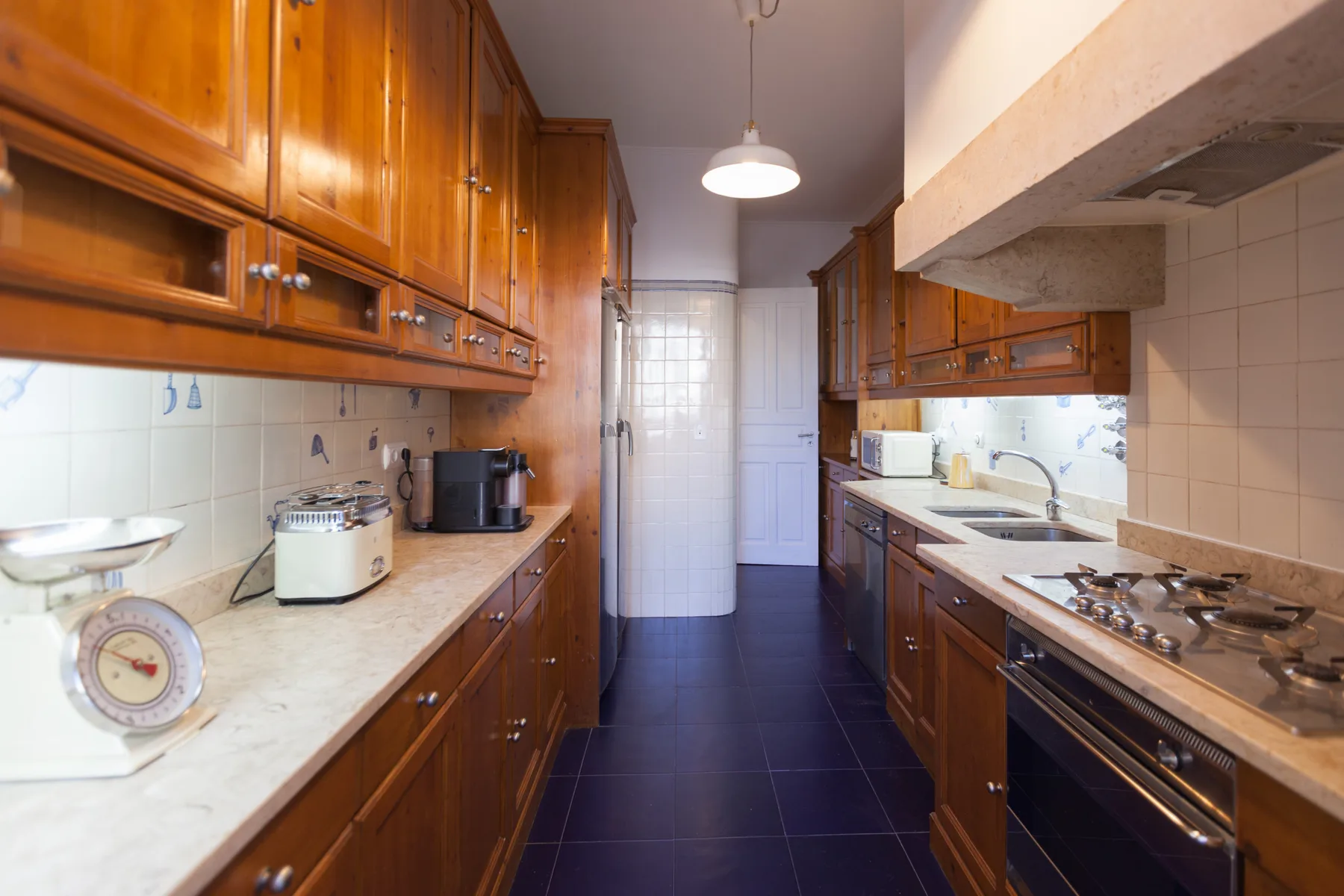 A galley kitchen with wooden cabinets, pale countertops, and dark floor tiles.