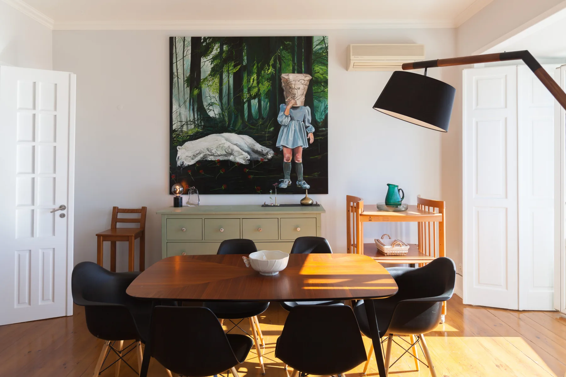 A dining area with a wooden table and chairs beneath a large forest painting.