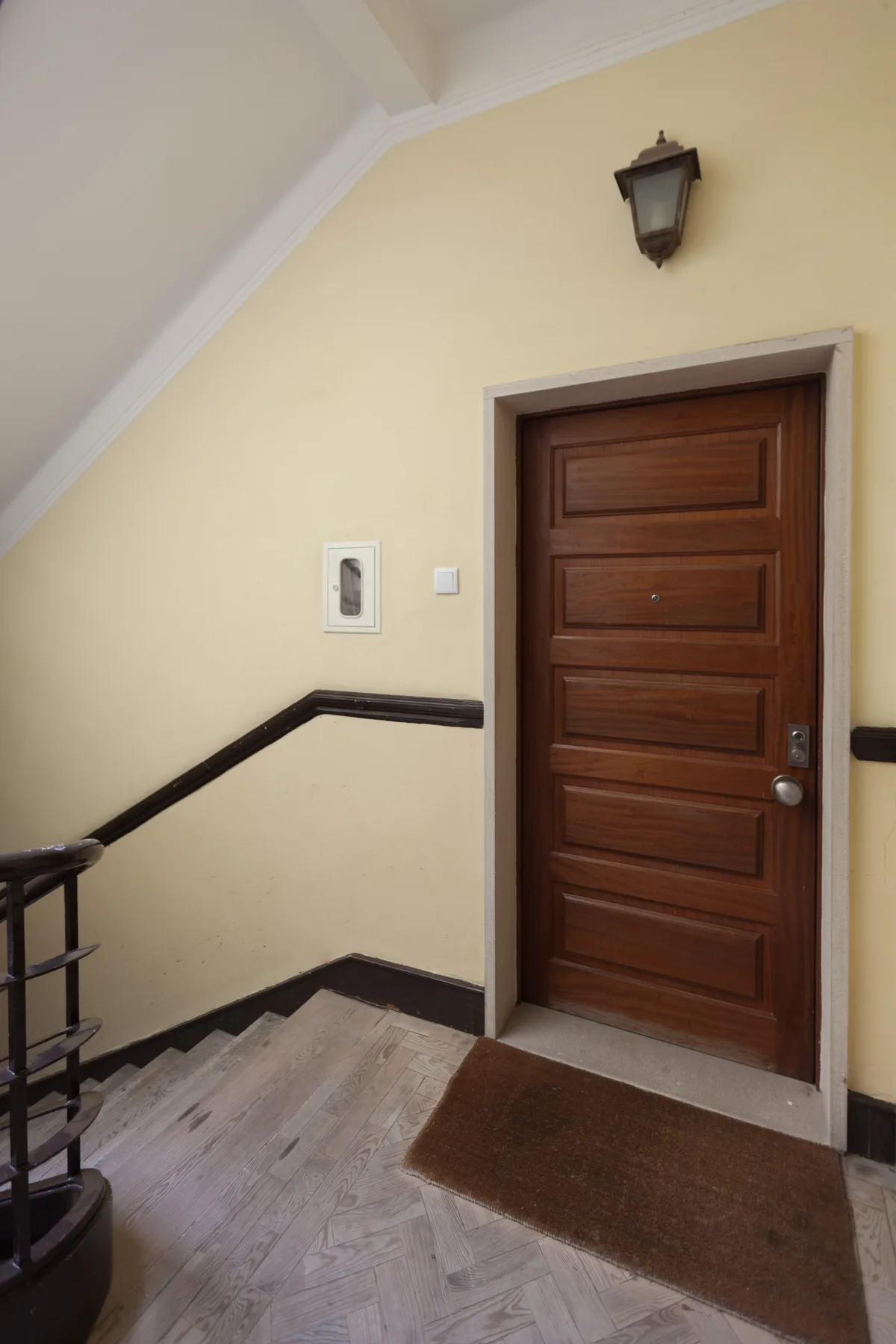 An apartment entry hall with a wooden door, wall light, and stair railing.