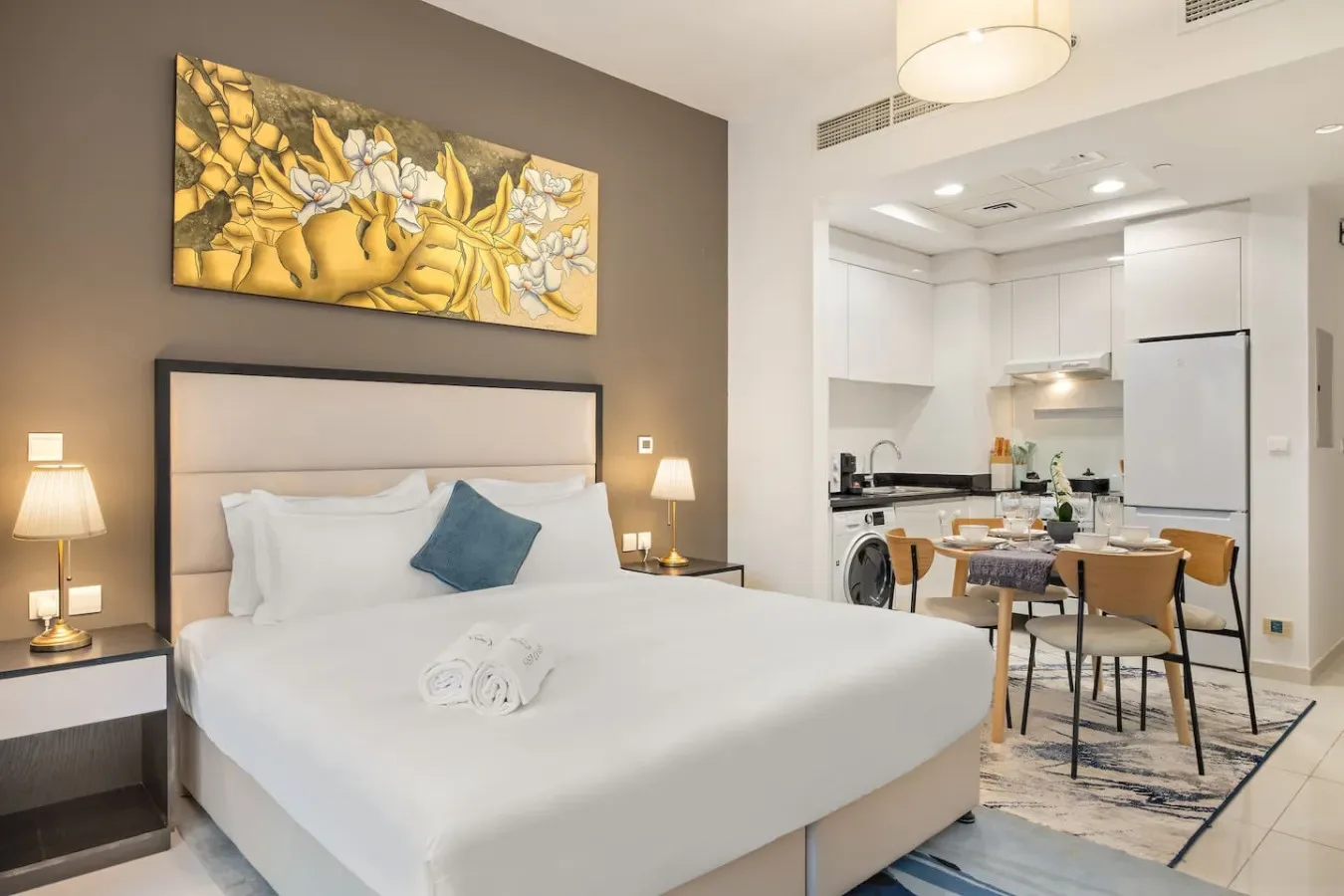 A bedroom with a neatly made bed opening onto a dining area and kitchen.