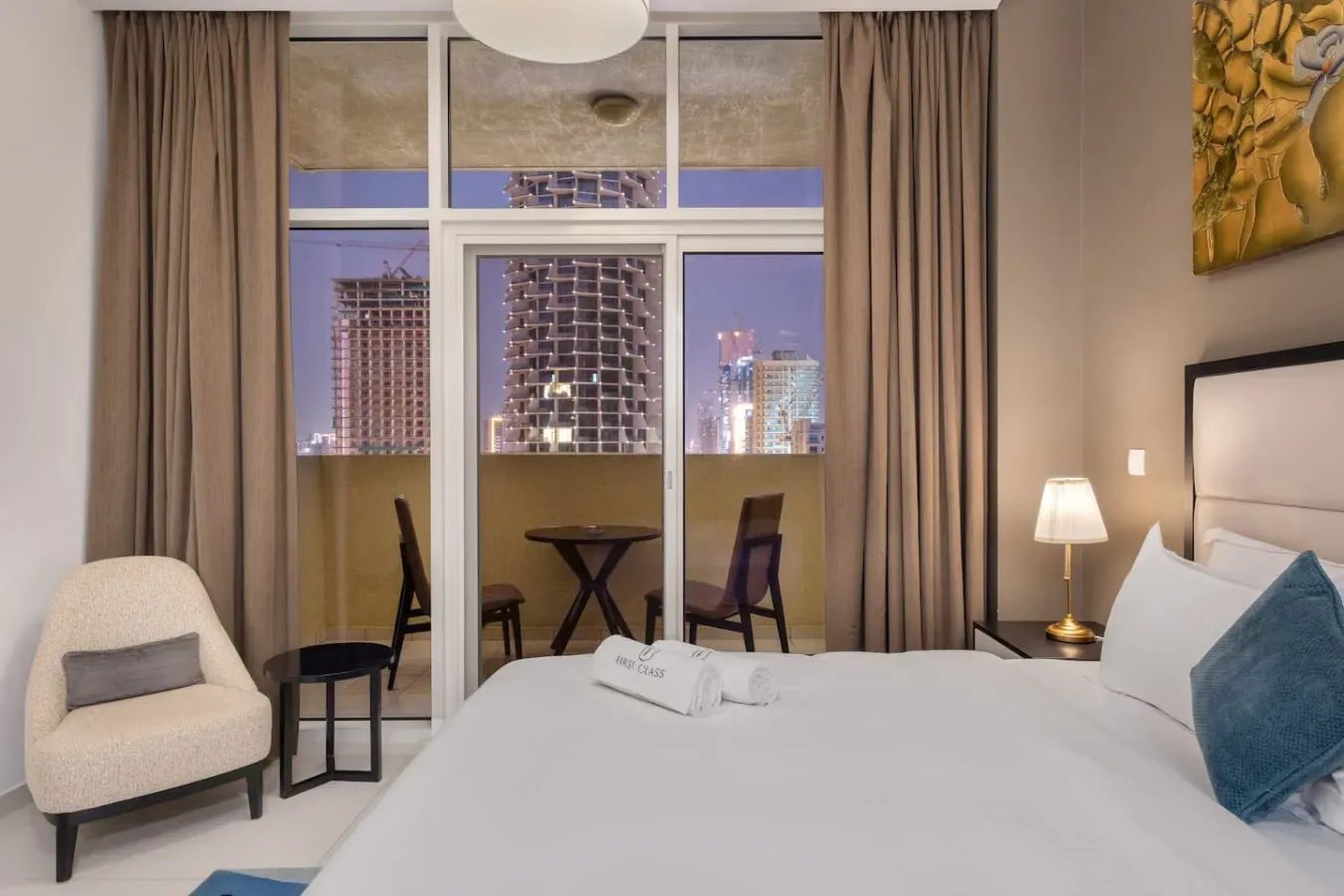 A bedroom with a bed, bedside lamp, and large windows overlooking city buildings.
