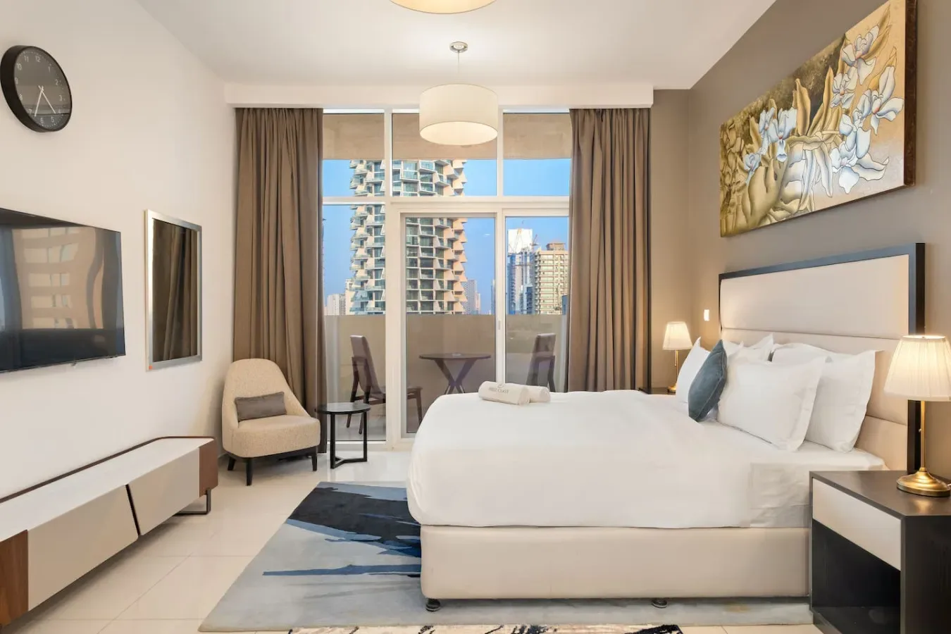 A bright bedroom with a bed, seating, and a large window overlooking nearby buildings.