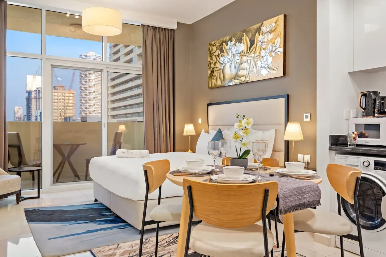 A bedroom with a bed and dining table beside large windows overlooking city buildings.