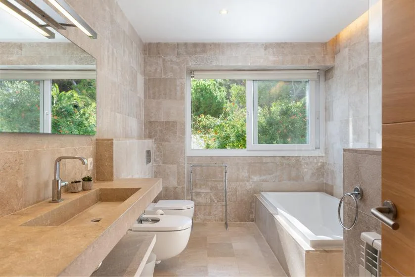A tiled bathroom with a bathtub, sink, and window overlooking greenery.