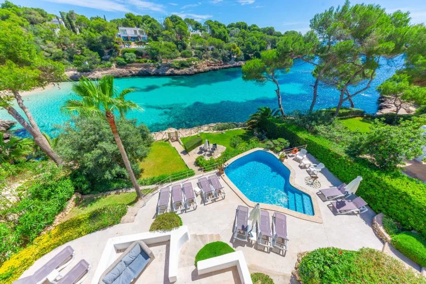 A swimming pool and sun loungers overlook a turquoise bay surrounded by greenery.