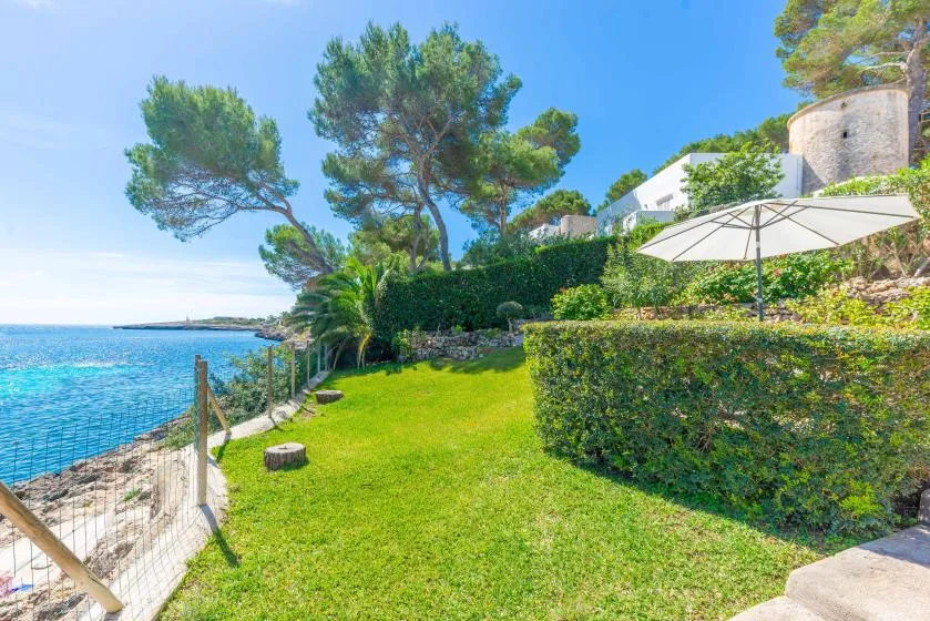 A grassy seaside garden overlooks blue water, with hedges and an umbrella nearby.