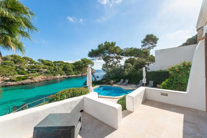 A sunlit terrace overlooks a turquoise cove, with a small pool nearby.