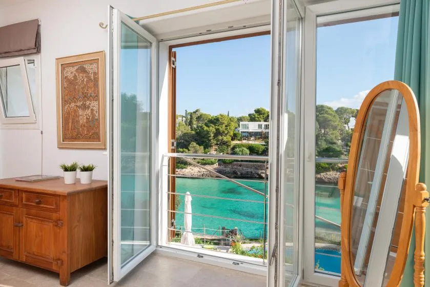 A bright room opens onto a balcony overlooking turquoise water and greenery.