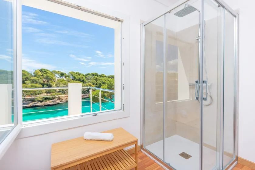 A bright bathroom with a glass shower and a large window overlooking turquoise water and greenery.