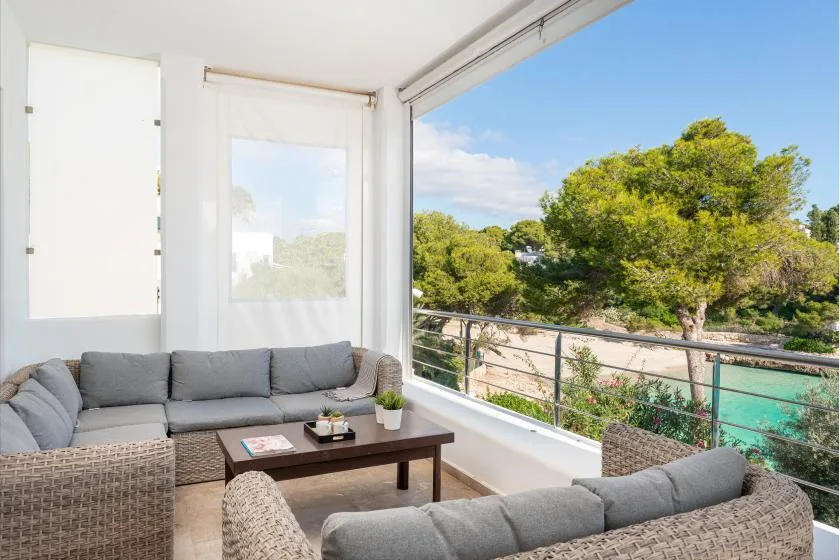 A bright living room opens onto a balcony overlooking trees and turquoise water.