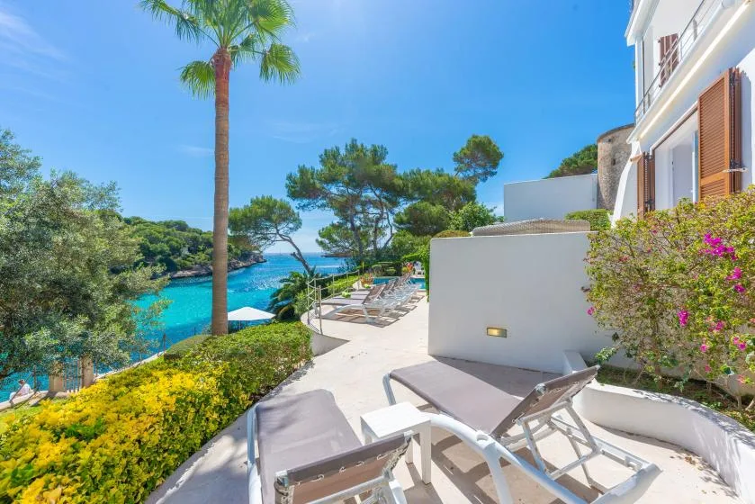 A sunny seaside terrace with reclining seating, greenery, and a view of the water.