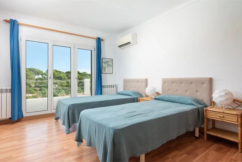 A bright bedroom with two beds and large glass doors opening onto a leafy view.