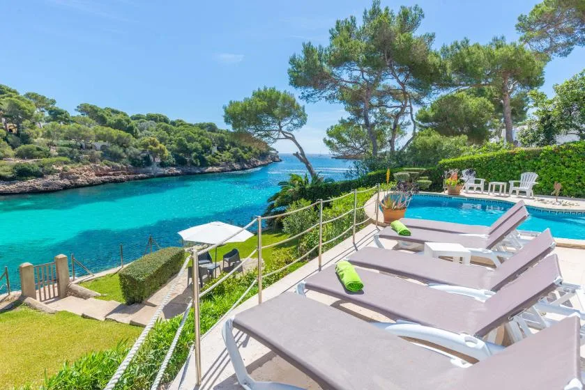 A poolside terrace overlooks turquoise water, with several loungers and pine trees.