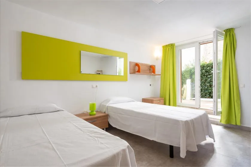 A bright bedroom with two beds and green curtains beside a glass door.