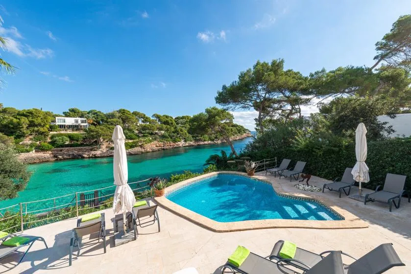 A swimming pool and sun loungers overlook a turquoise coastal inlet.