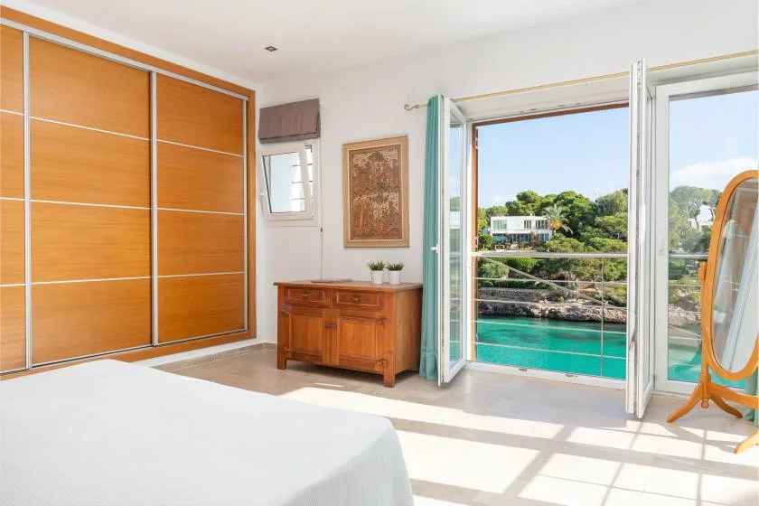 A bright bedroom opens onto a balcony overlooking turquoise water and greenery.