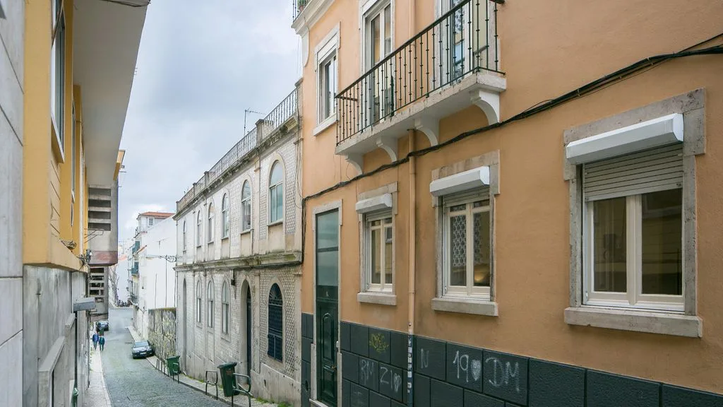 Una calle estrecha pasa entre edificios cercanos con balcones y ventanas con persianas.